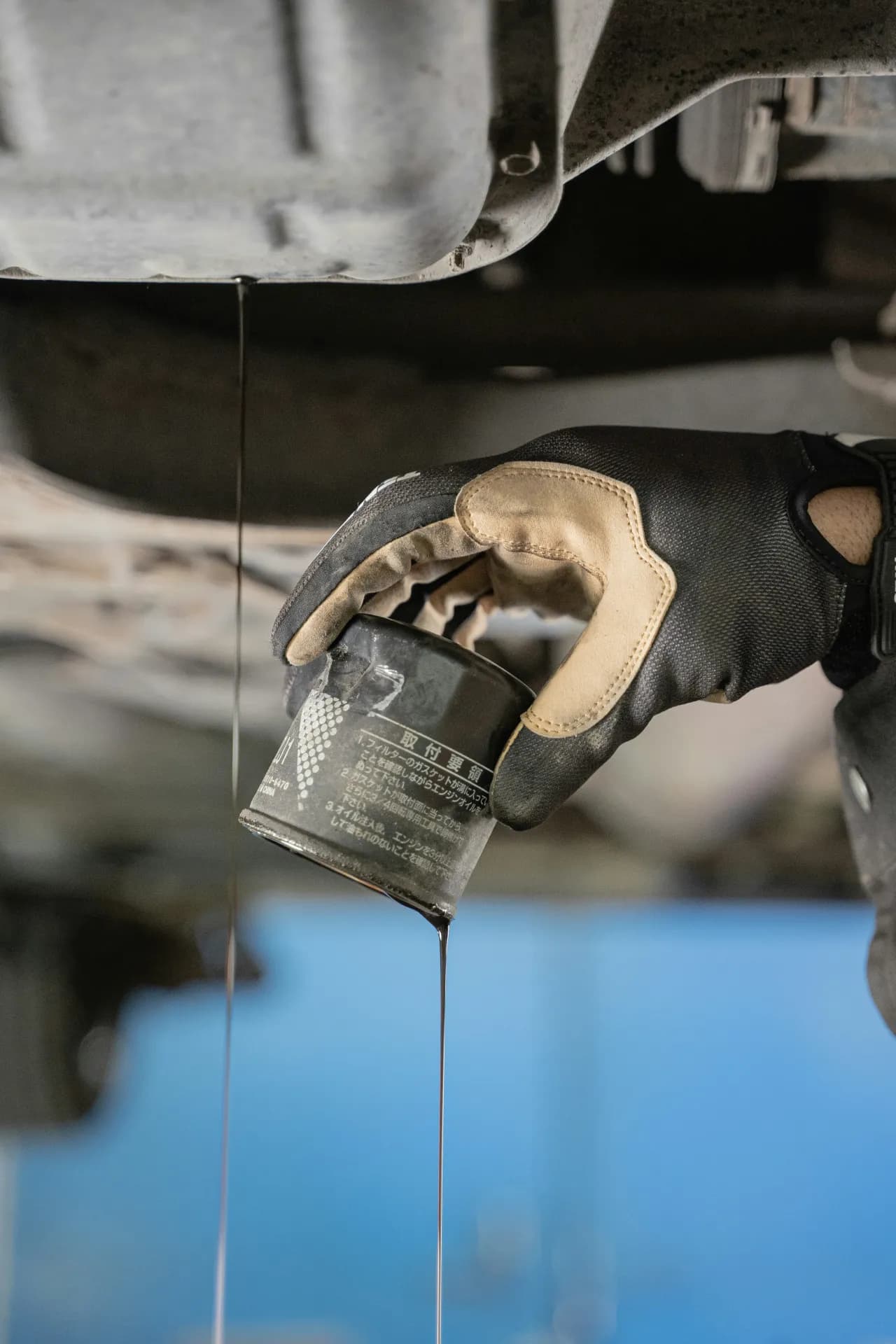 Hand in einem Arbeitshandschuh hält einen Ölfilter unter ein Fahrzeug, dunkles Öl tropft herab