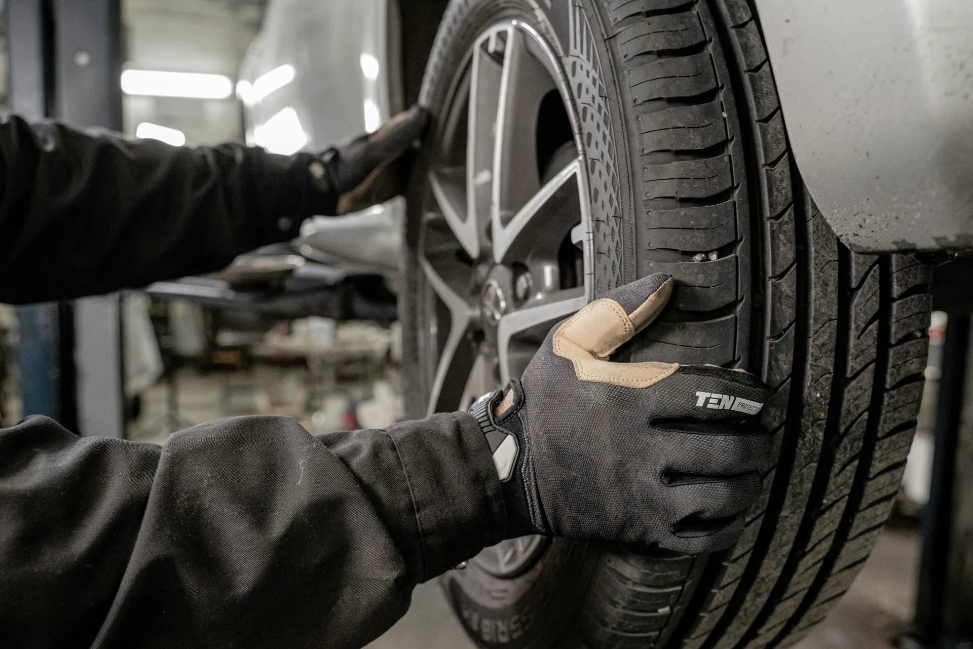 Zwei Hände in Arbeitshandschuhen halten einen Reifen mit Leichtmetallfelge an einem Fahrzeug in der Werkstatt